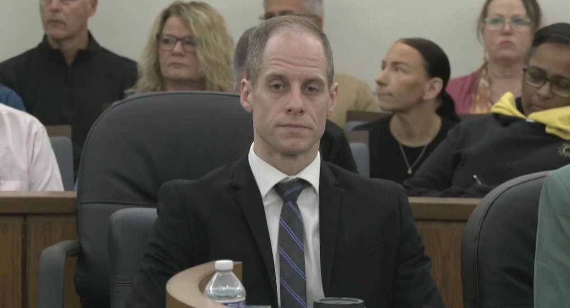 A man in a dark suit and striped tie sits at the defense table in a courtroom, with spectators seated in the gallery behind him