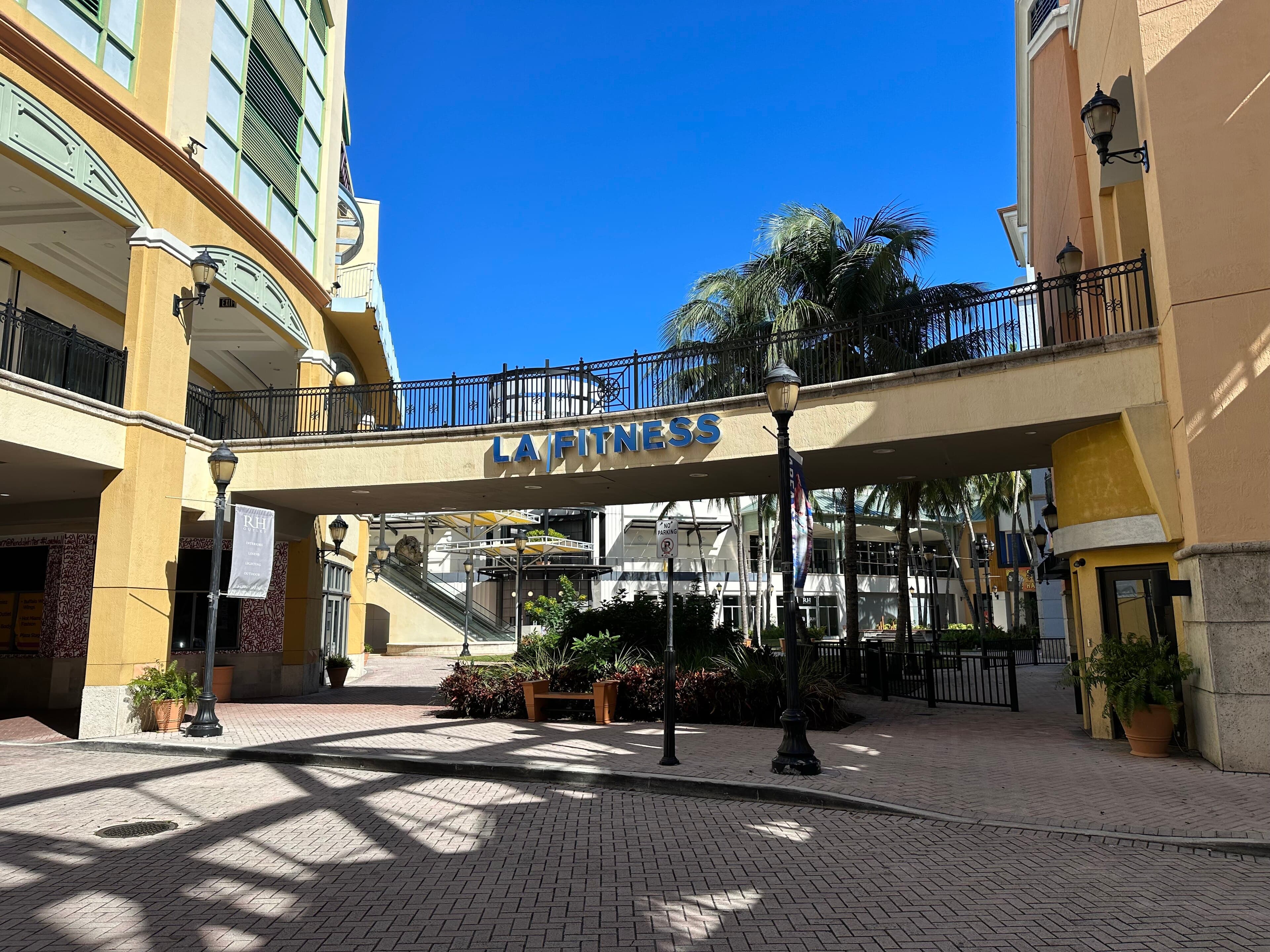 An empty brick walkway at the Shops at Sunset Place, with a pedestrian bridge overhead and palm trees under a blue sky