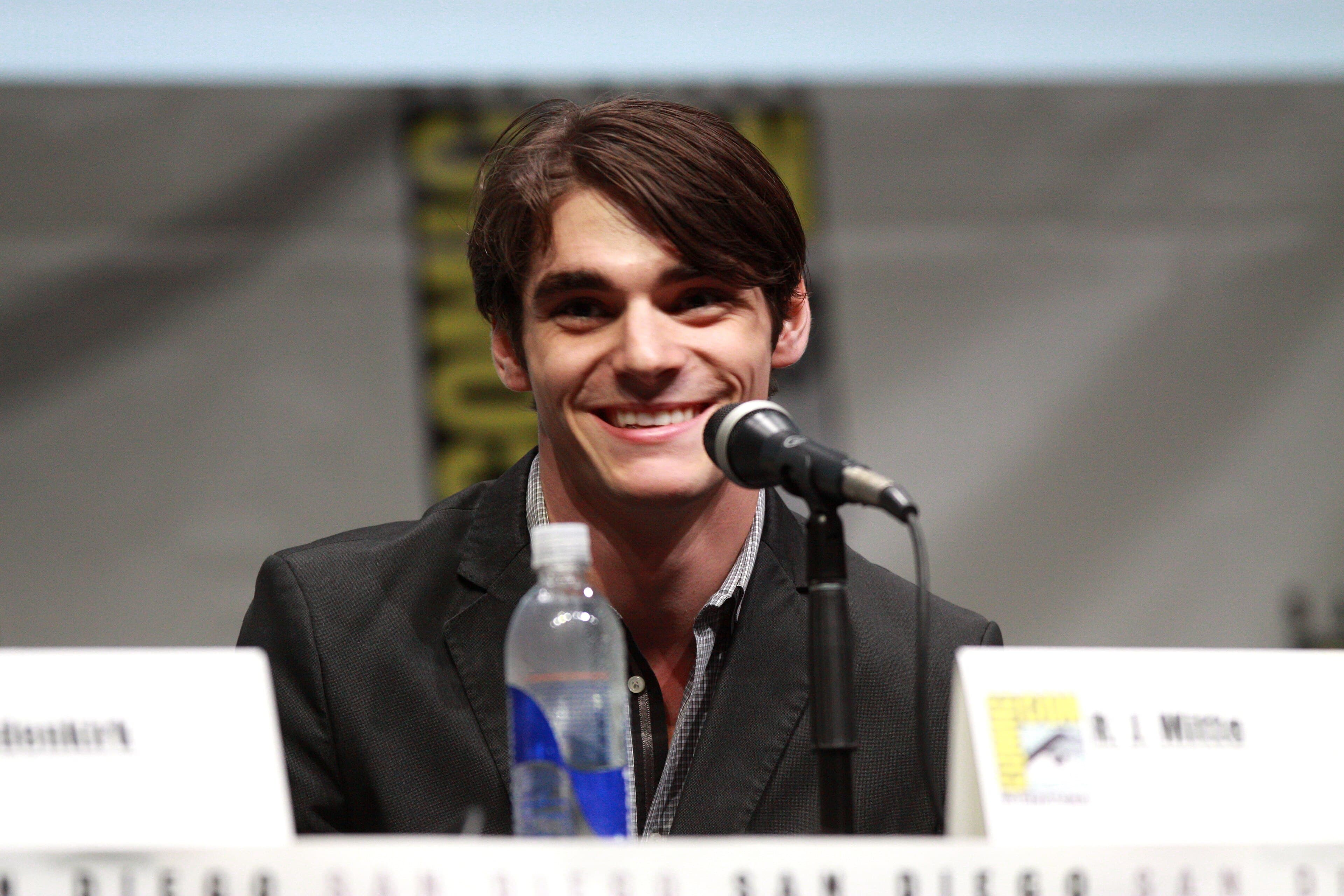 RJ Mitte smiles while speaking into a microphone at a panel table.