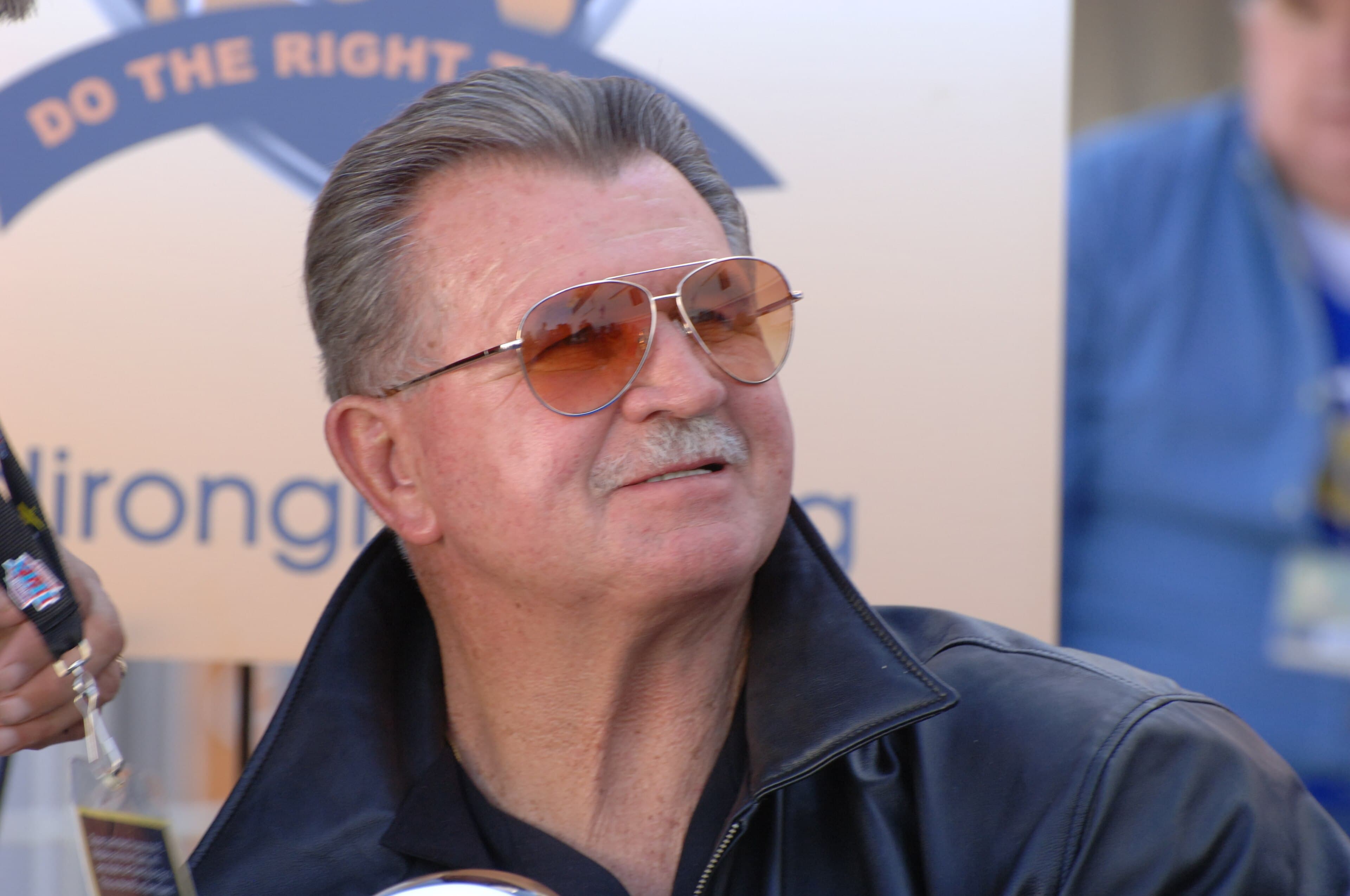 Mike Ditka, with gray hair, mustache and tinted aviator sunglasses, smiling at an event