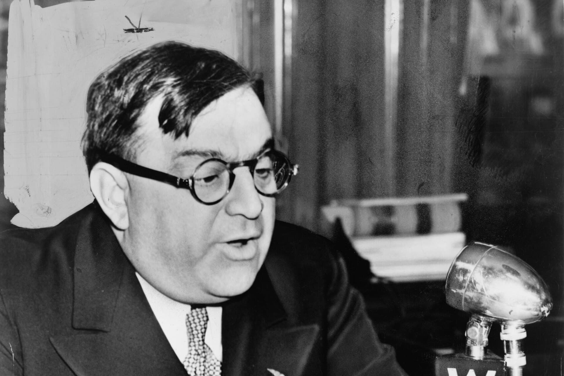 Black-and-white photo of Fiorello La Guardia in round glasses, reading from papers at a desk microphone labeled WNYC