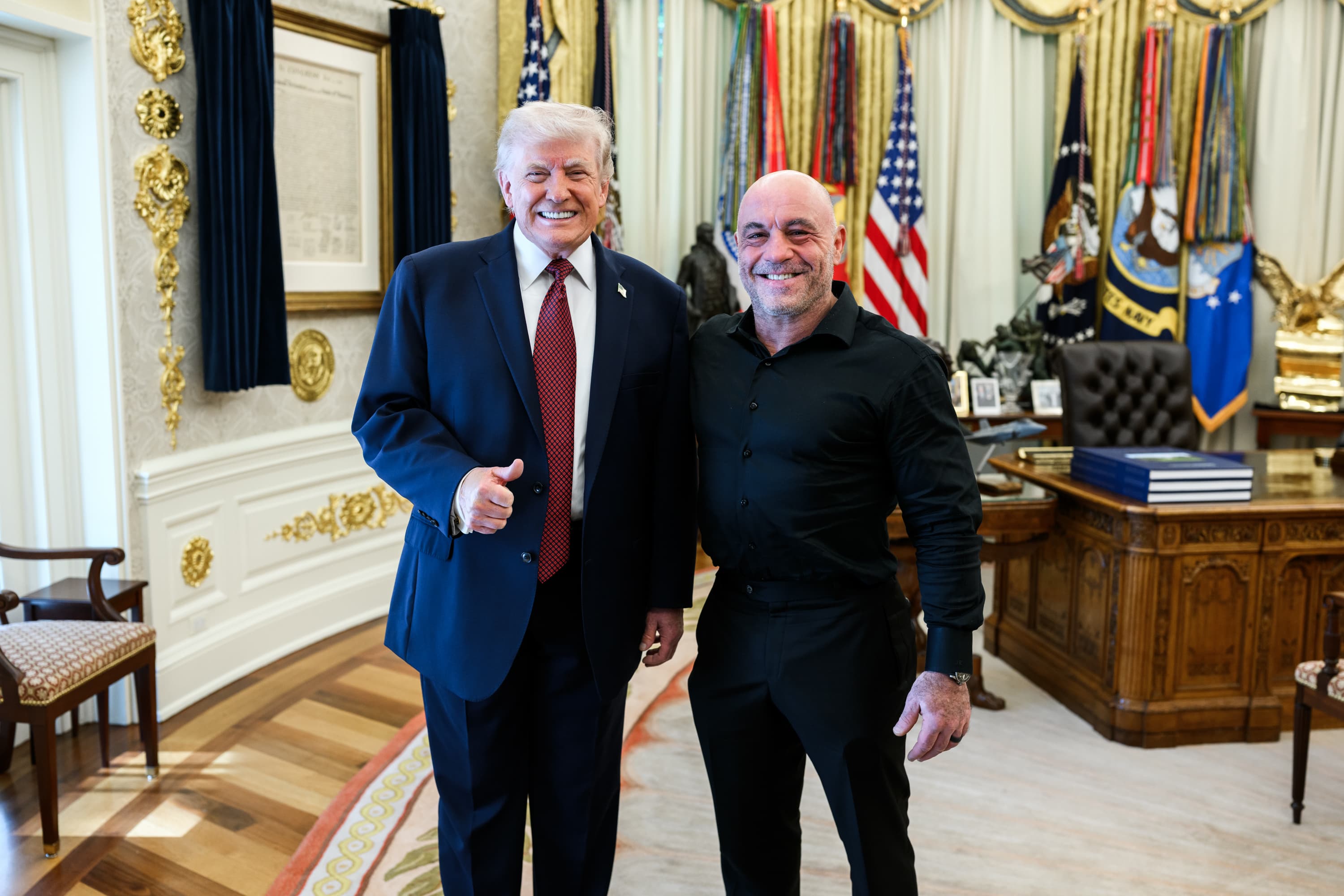 Joe Rogan, in black, stands smiling beside President Donald Trump, who gives a thumbs-up, in the Oval Office