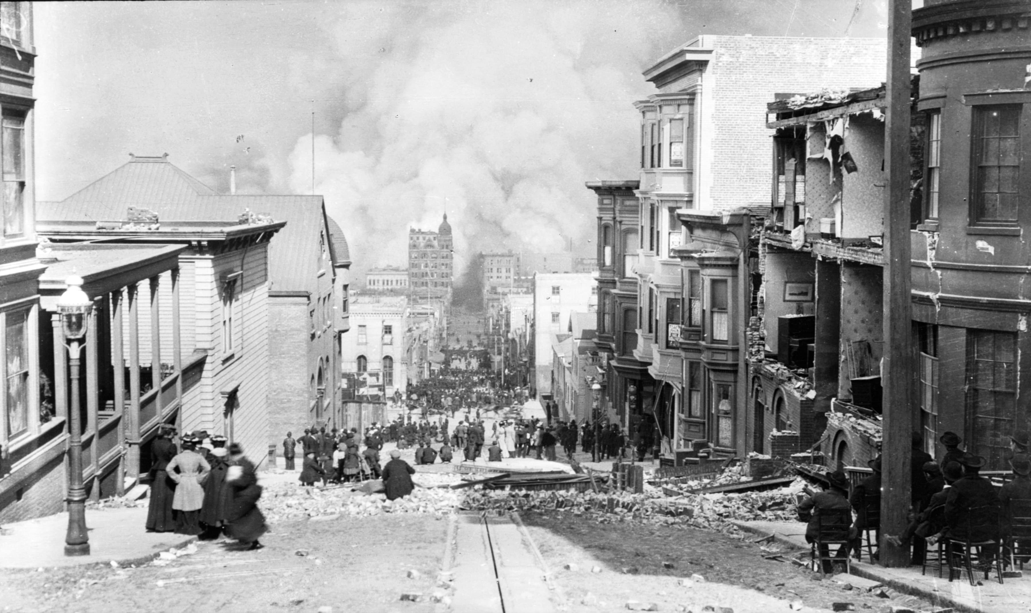 Black-and-white photo looking down a steep San Francisco street toward billowing smoke from the 1906 fire, with onlookers standing in the road