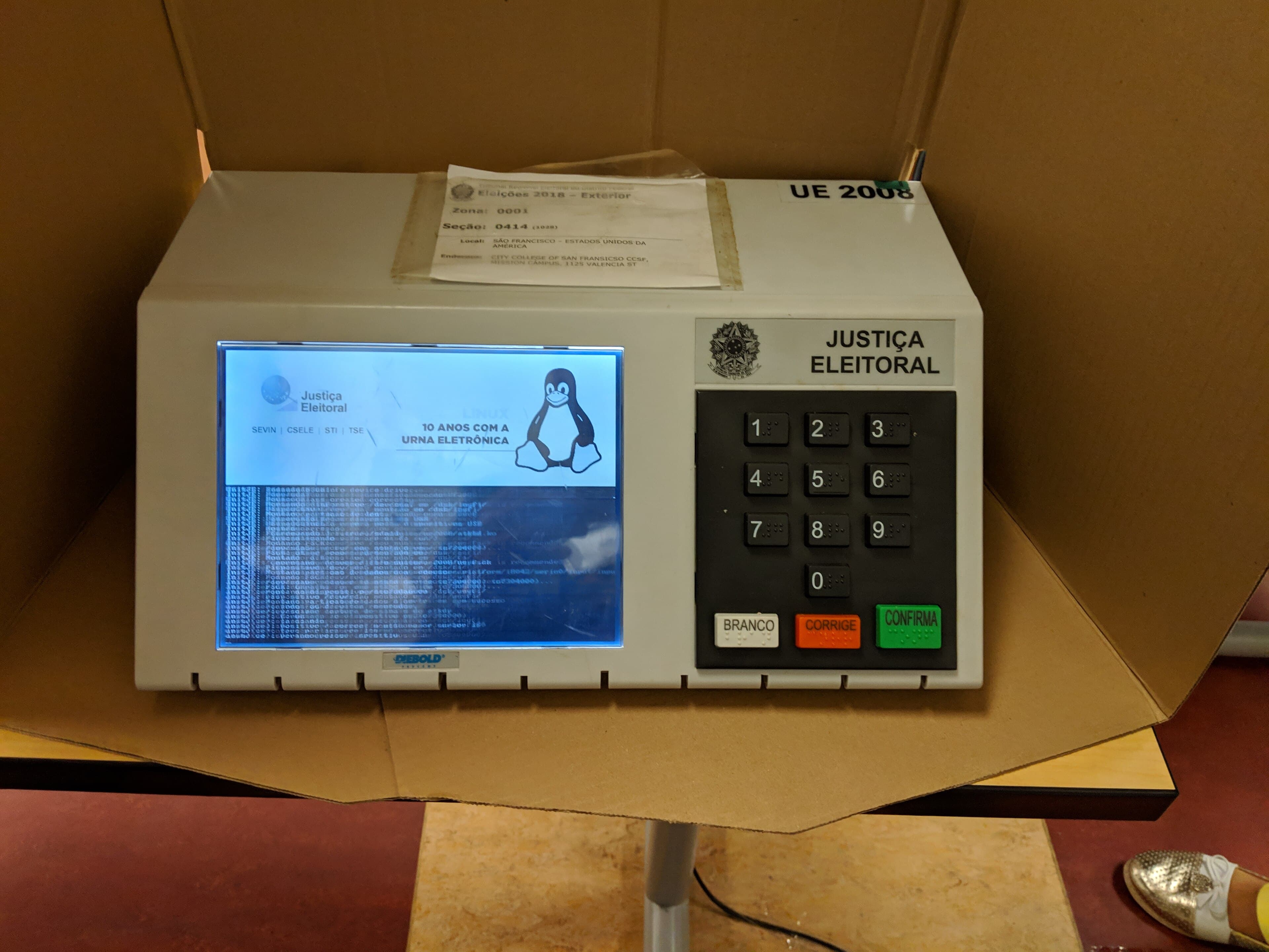 A Brazilian electronic voting machine inside a cardboard voting booth, its screen lit as it starts up