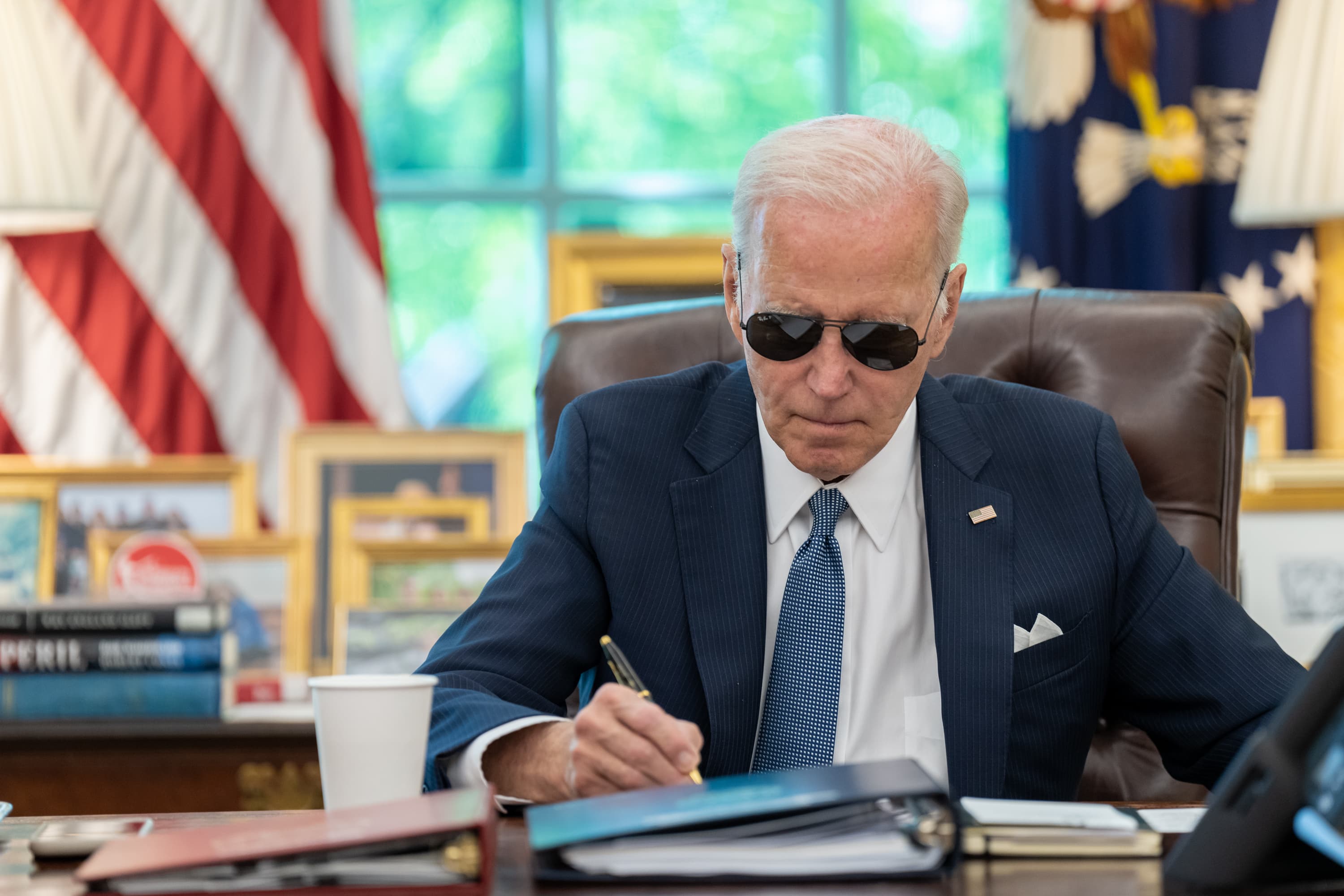 Joe Biden in aviator sunglasses signing papers at the Resolute Desk in the Oval Office