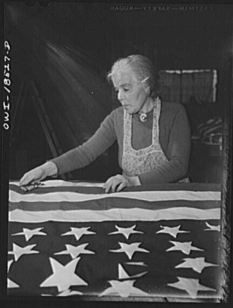 Black-and-white photo of an older woman in an apron sewing a large American flag at a table