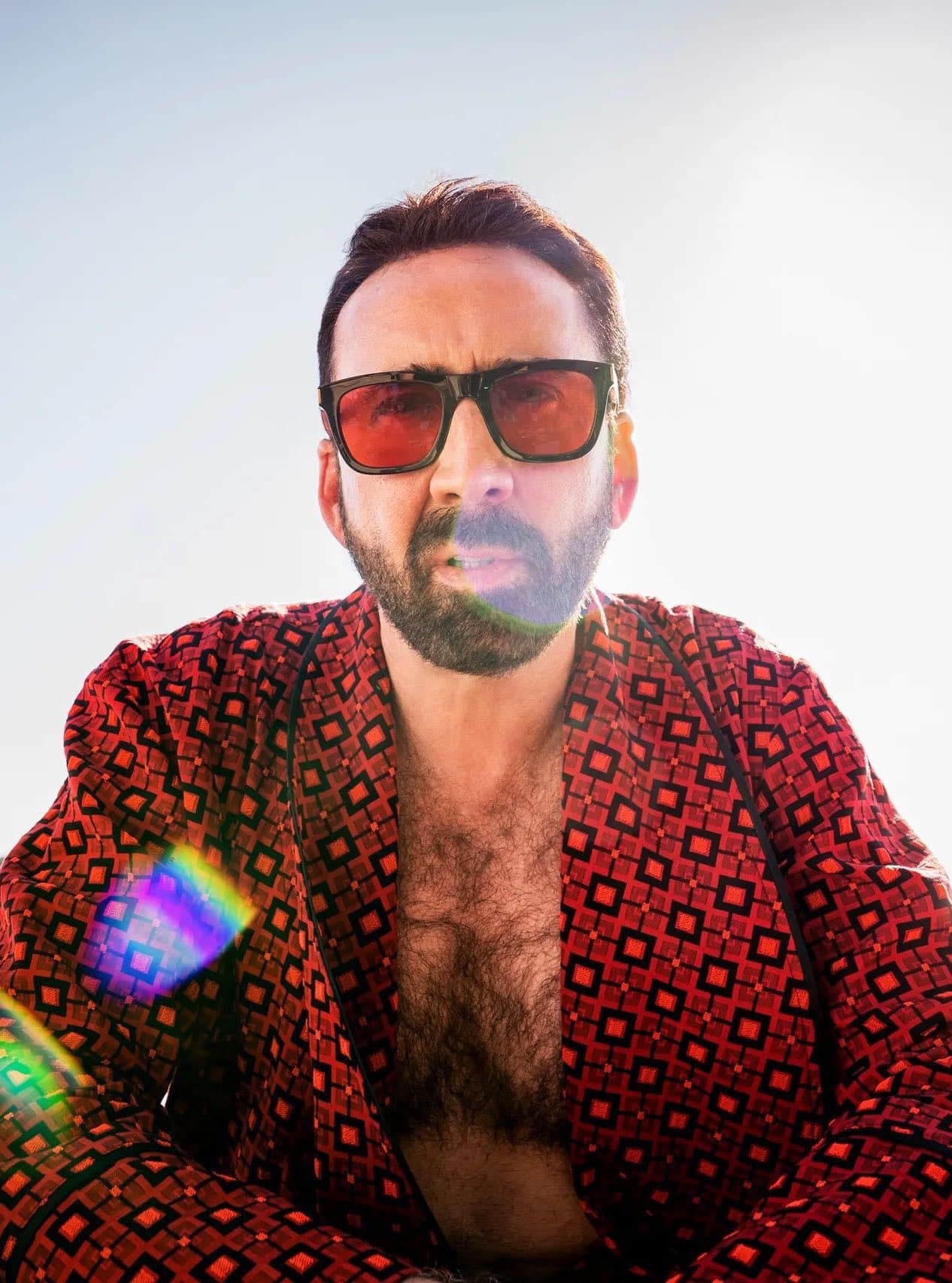 Nicolas Cage in red-tinted sunglasses and an open red patterned robe against a bright sky