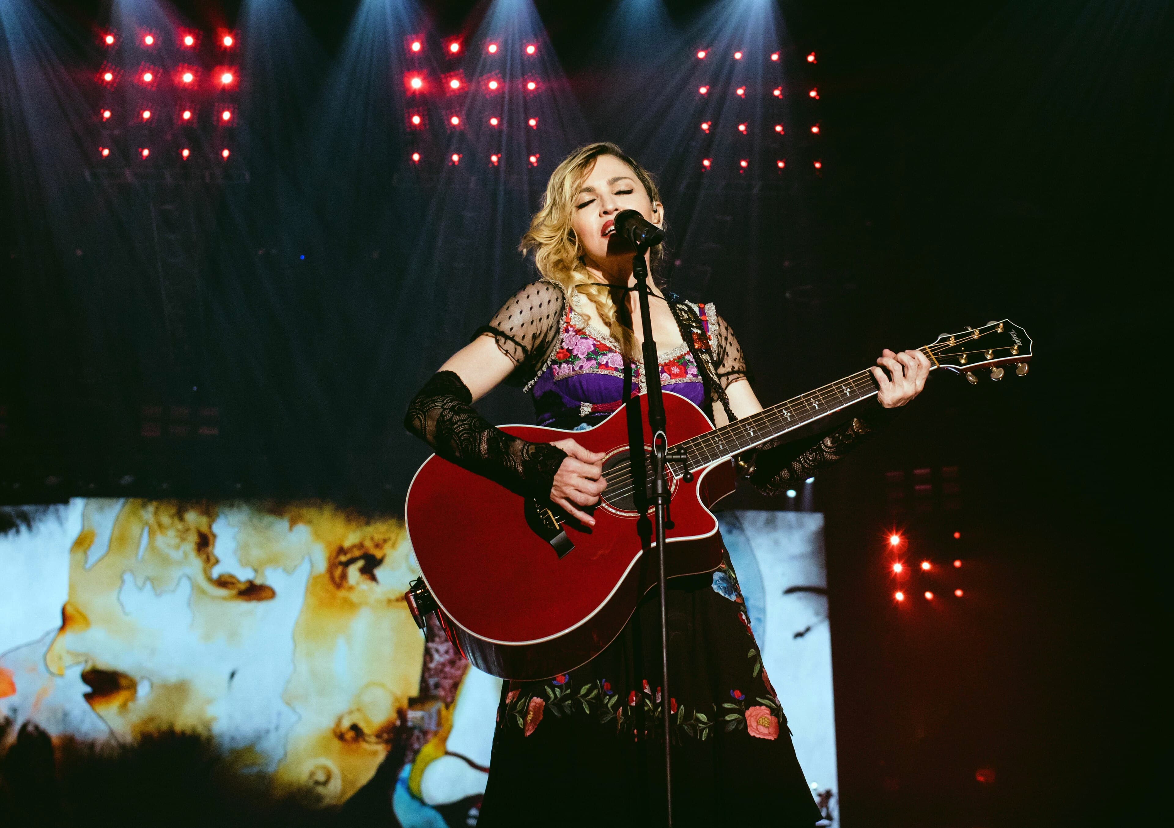 Madonna singing into a microphone and playing a red acoustic guitar on stage under red lights